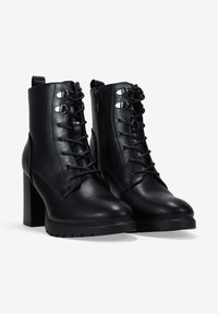 Botas de tobillo de cuero negro con puntera afilada, cierre de cordones en la parte delantera, acentos de hardware metálico y un tacón alto y robusto. Presenta suelas texturizadas.