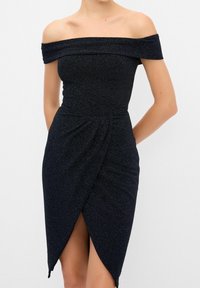 Femme portant une robe noire ajustée, épaules dénudées, en tissu texturé avec une fente à l'avant, posant devant un fond uni.