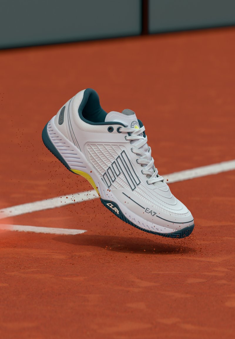 Fehér atlétikai cipő texturált mintázattal, sötétkék díszítéssel és sárga részlettel a középtalpán, levegőben, egy salakpályán.