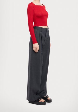 Kvinde iført tætsiddende rød crop top med lange ærmer, højtaljede, brede grå bukser og sorte platformsandaler med åbne tæer.