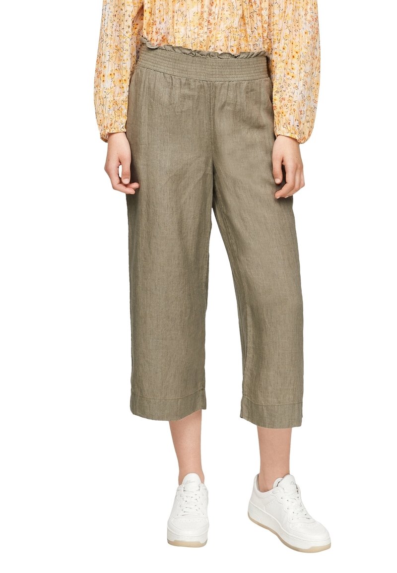 Femme portant un pantalon large court vert olive, un chemisier fleuri à manches longues et des baskets blanches, debout devant un fond blanc.