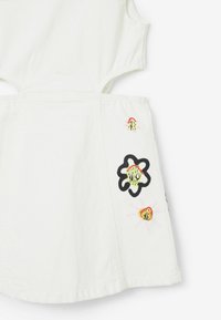 Bílá bezramínková šaty s květinovými vzory v zelené, růžové a černé barvě. Obsahuje výřezy po stranách a texturovanou tkaninu.