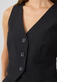 Crna bezrukavna prsluk s V izrezom, zatvaranje na tri gumba i dva bočna džepa; izrađen od glatkog materijala s krojem koji prati tijelo.