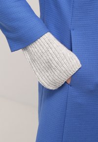 Blue textured fabric with ribbed gray knit sleeve. Sleeve extends from a fitted coat with side pockets. Focus on shape and material contrasts.