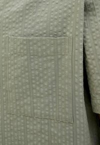 Green textured fabric with a grid pattern, featuring a patch pocket. Material appears soft, with visible stitching details along the edges.