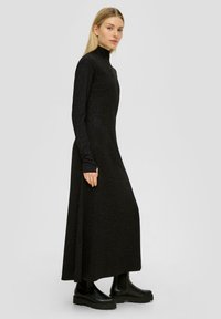 Longue robe noire avec un col montant, des manches longues et une coupe ample. Fabriquée en tissu texturé avec un léger éclat. Assortie à des bottes noires épaisses.