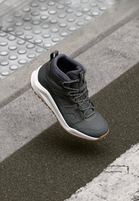 Scarpa da trekking grigia con una tomaia testurizzata, tallone rinforzato e lacci riflettenti. Presenta un intersuola bianca e una suola in gomma marrone per una buona aderenza.