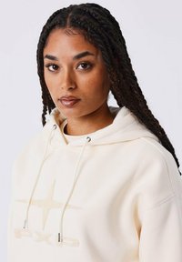 Jonge vrouw met gevlochten haar, gekleed in een crèmekleurige hoodie met een gestructureerde ster en het "PXG"-logo aan de voorkant, kijkend naar voren tegen een effen achtergrond.