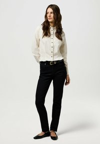 Camisa blanca de botones con un patrón texturizado, combinada con jeans negros ajustados. Bailarinas negras con tiras al tobillo completan el atuendo.