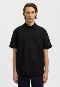 Jeune homme aux cheveux bruns mi-longs, portant une chemise noire à manches courtes et boutons, debout devant un fond blanc.