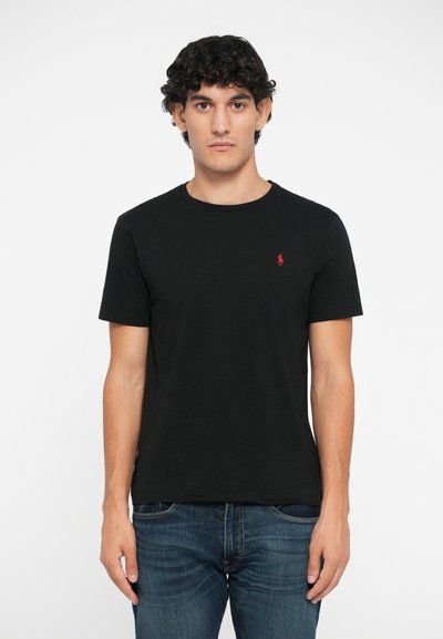 Camiseta de algodón negra con cuello redondo, mangas cortas y un pequeño logo rojo en el lado izquierdo del pecho. Combinada con pantalones vaqueros de mezclilla azules.