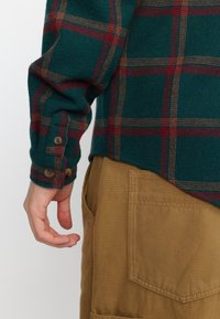 Groene en rode geruite flanellen overhemd met een knoopmanchet, gecombineerd met tan stoffen broeken met een zijzak. Geverfd wolmateriaal.