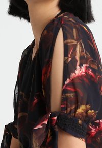 Průhledná černá květinová blůza s krátkými rukávy a výřezem, ozdobená stuhami a krajkovým lemem, která vykazuje živý květinový vzor.