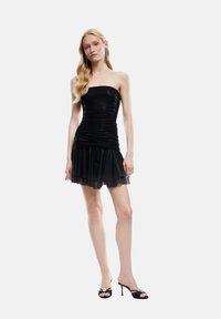 Schwarzes trägerloses Kleid mit geraffter Textur. Der kurze Rock besteht aus geschichtetem Tüll. Das Model trägt schwarze Open-Toe-Absätze mit Perlenakzenten.