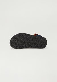 Crna gumena potplat s valovitim uzorkom; sadrži narančastu i plavu traku smještenu straga. Ravni profil, bez ukrasa.