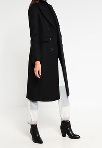 Sort uldfrakke med en lang silhuet, enkeltknap, notch lapels og to forlommer, parret med sorte ankelstøvler og grå bukser.