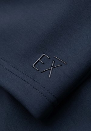 Dunkelblauer Stoff mit Nahaufnahme von genähten Nähten und einem erhabenen, glänzenden "EX" Logo-Detail auf dem Material.