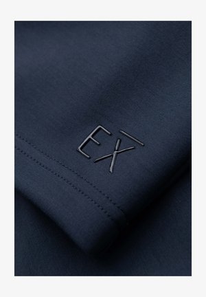 Dunkelblauer Stoff mit Nahaufnahme von genähten Nähten und einem erhabenen, glänzenden "EX" Logo-Detail auf dem Material.