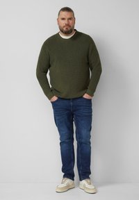 Oliwkowy dzianinowy sweter z okrągłym dekoltem, zestawiony z niebieskimi dżinsami i kremowymi sneakersami z brązowymi akcentami.