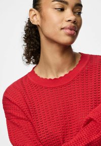 Maglione rosso, con texture e scollo a scallop, realizzato con un motivo a maglia larga. Presenta maniche lunghe, polsini a costine e un design dalla vestibilità rilassata.