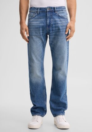 Homme portant un jean bleu moyen à jambes droites et des baskets blanches, debout devant un fond clair uni.