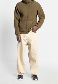 Felpa zip-up marrone con colletto alto, pantaloni di cotone crema e scarpe da ginnastica nere con dettagli viola.