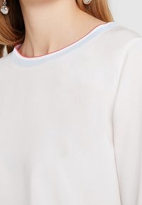 Top bianco con scollo a giro colmo e bordi rossi e azzurri chiari. Texture liscia e design minimal con dettagli di cucitura visibili.