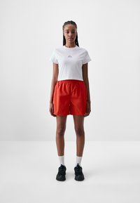 White cropped t-shirt with blue logo, paired with red shorts featuring an elastic waistband. Model wears black sneakers and white socks.