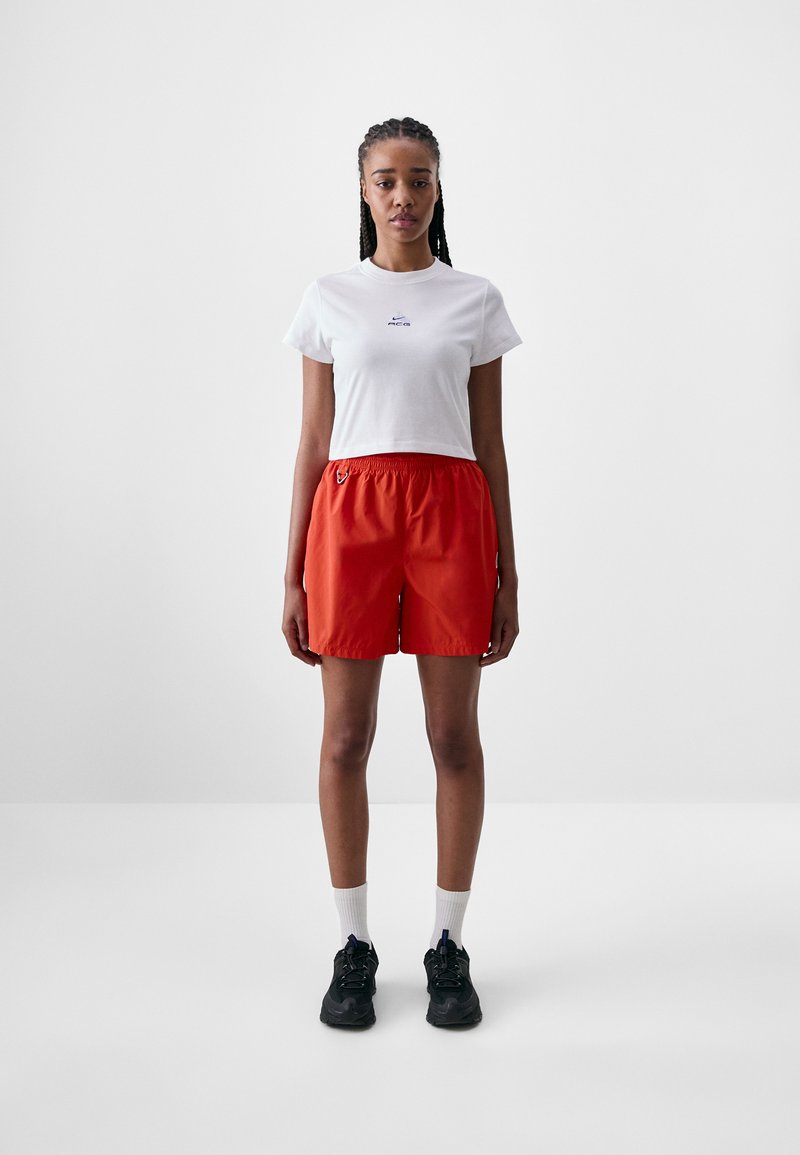 White cropped t-shirt with blue logo, paired with red shorts featuring an elastic waistband. Model wears black sneakers and white socks.