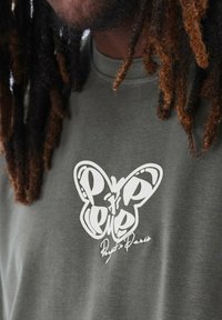 Camiseta verde oliva con un gráfico de mariposa blanca y diseño de texto. Hecha de algodón suave, con cuello redondo y mangas cortas.