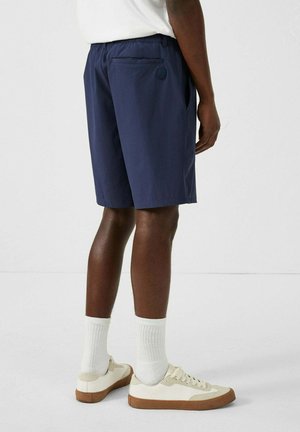 Marineblauwe shorts met een relaxed fit, elastische tailleband en achterzak; gecombineerd met witte sneakers en sokken. Minimalistisch ontwerp en een soepele textuur.