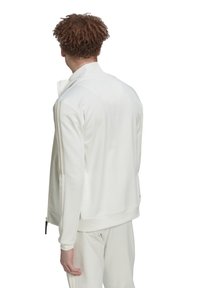 Vit zip-up jacka i mjukt tyg, med hög krage, ribbad fåll och subtila sidostråk. Baksidan visar en figurnära design.