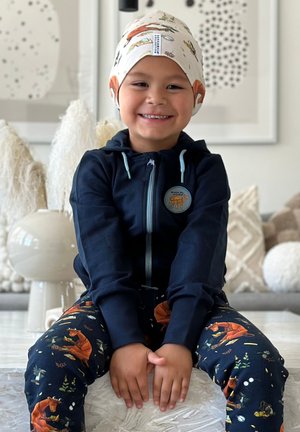 Barn iført en marineblå zip-up hoodie med en rund applikation og mønstrede pyjamasbukser med orange ræve og planter. Stoffet er blødt og afslappet.