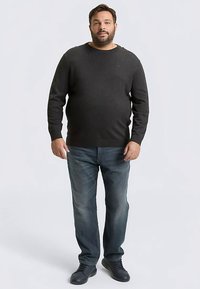 Mörkgrå stickad tröja med rund halsringning, kombinerad med blekta blå jeans och svarta sneakers, stående mot en ljus bakgrund.