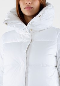 Giacca puffer bianca con colletto alto, chiusura con cerniera e dettagli dorati. Tessuto leggero e testurizzato con cuciture orizzontali.