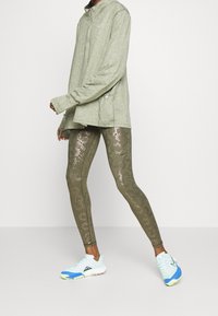 Jasnozielona bluzka z długim rękawem, wzorzyste oliwkowe legginsy z metalicznymi kwiatowymi akcentami oraz białe sneakersy z niebieskimi i zielonymi detalami.