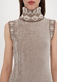 Haut sans manches en tricot beige clair et crème. Il présente un col montant à côtes avec des motifs géométriques et des rayures texturées sur l'ensemble.