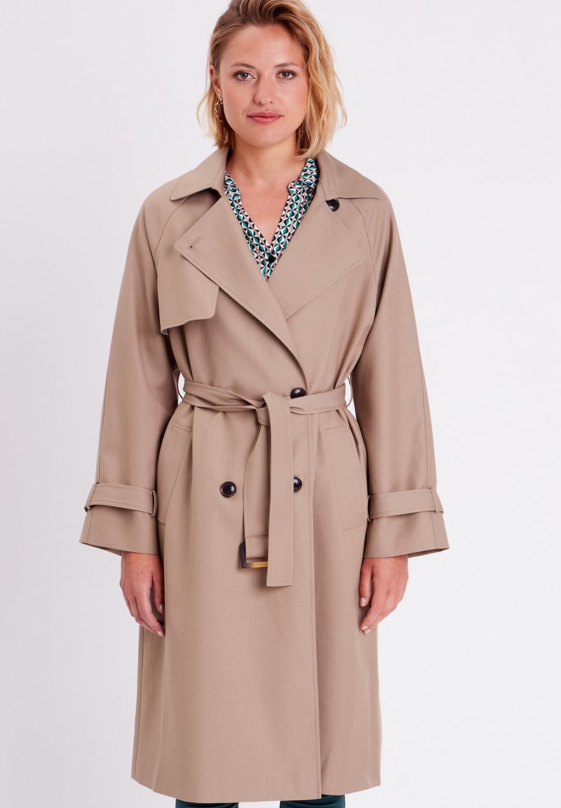 Gabardina beige con un frente de doble botonadura, solapas anchas y puños enrollados. Presenta un cinturón a juego y pequeños adornos de botones.