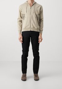 Beige Kapuzenpullover mit Reißverschluss und Kordelzug, versehen mit schwarzen Engel-Grafiken auf der Brust, kombiniert mit schwarzen Slim-Fit-Jeans und braunen Sneakers.
