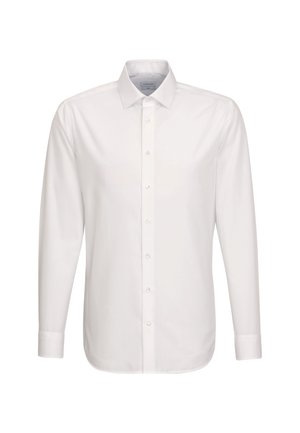 Un jeune homme se tient face à l'objectif, vêtu d'une chemise blanche à boutons, d'un pantalon plissé foncé avec des revers, de chaussures noires et de chaussettes foncées, sur un fond uni.