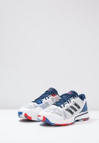 Scarpe sportive bianche con accenti blu, tomaia in rete, colletto imbottito e dettagli rossi sulla suola. Tre strisce nere sui lati.