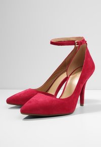 Scarpe rosse in suede con tacco alto, punta affusolata e cinturino regolabile alla caviglia. Texture liscia, interno leggermente imbottito e tacco a stiletto sottile.
