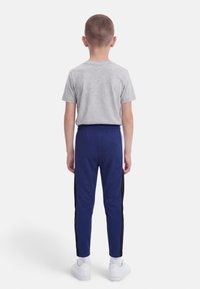 Παιδί που στρέφεται προς το μέρος του, φοράει ανοιχτό γκρι κοντομάνικη μπλούζα, μπλε φόρμα με μαύρες ρίγες στο πλάι, λευκές κάλτσες και λευκά αθλητικά παπούτσια.