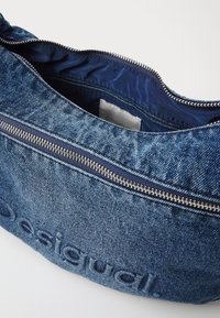 Dżinsowa torba z zamknięciem na zamek, ciemnoniebieska podszewka, wytłoczone logo na zewnętrznej stronie oraz teksturowana tkanina.