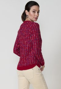 Pull en tricot rouge avec des accents bleus et blancs, présentant un motif texturé, un col rond et des poignets côtelés, associé à un pantalon de couleur crème.