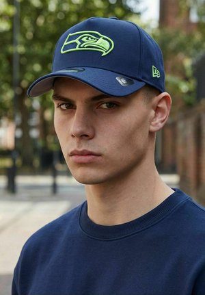 Young man outdoors wearing a navy blue cap with a green hawk logo and a matching navy sweatshirt, looking directly at the camera.