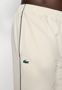 Pantaloni corti color crema realizzati in tessuto leggero, con superficie testurizzata, vita elasticizzata e logo di coccodrillo ricamato in verde sul lato.