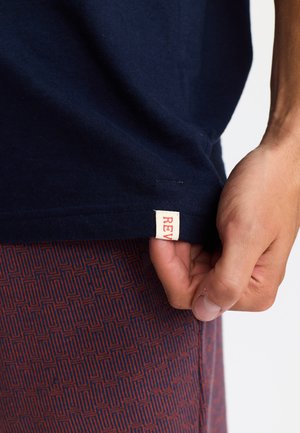 T-shirt en coton bleu marine avec une étiquette crème visible portant du texte rouge. Une main saisissant l'ourlet. Des shorts à motifs légers visibles en dessous.