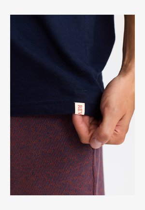 T-shirt en coton bleu marine avec une étiquette crème visible portant du texte rouge. Une main saisissant l'ourlet. Des shorts à motifs légers visibles en dessous.