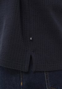 Tissu en maille gaufrée bleu marine foncé avec une petite étiquette noire de taille indiquant « M » au-dessus d'une fente sur l'ourlet, par-dessus un denim bleu foncé avec des surpiqûres orange.
